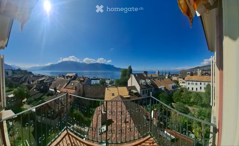 Balcony overlooking a town with a lake and mountains in the background, featuring a metal railing and wooden decking.
