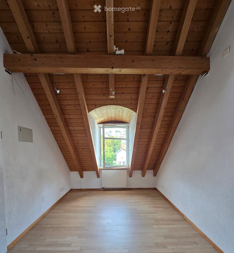 Empty room with wooden floor, exposed wooden ceiling beams, white walls, and a large arched window letting in natural light.