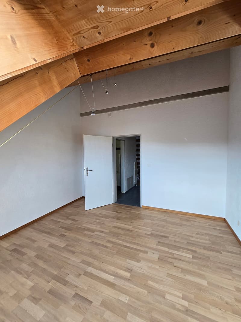 Empty room with light wooden flooring, white walls, and a white door leading to another space. Exposed wooden beams on the ceiling with hanging lights.