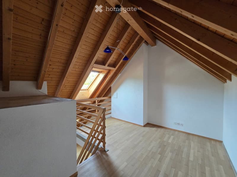 Bright, empty attic space with wooden beams, skylight windows, and a wooden staircase leading down. Light blue walls and wooden flooring.