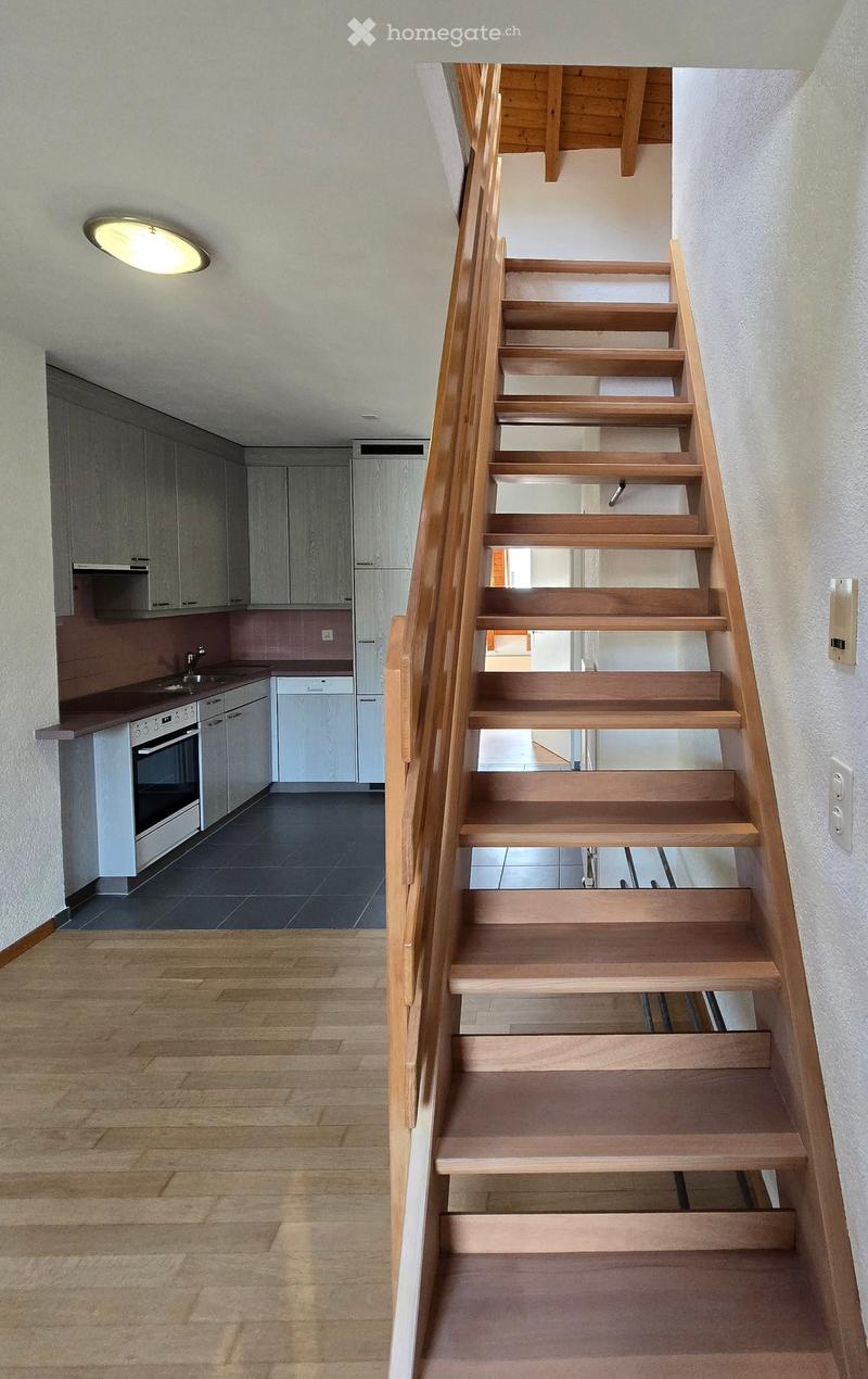 Wooden staircase with a modern design, leading to a kitchen area with white cabinets, stainless steel appliances, and a dark countertop.
