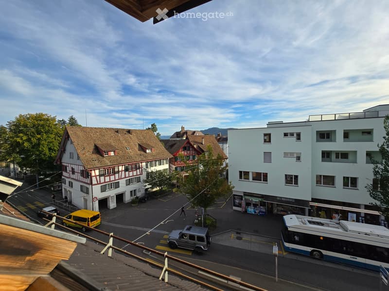 View of a town square with traditional houses, modern buildings, and vehicles including a bus and a van.