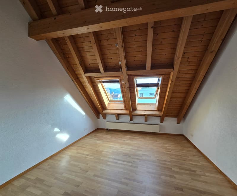 Empty attic room with wooden floors, exposed wooden beams, and two skylight windows. The walls are painted white, and there is a radiator on one wall.