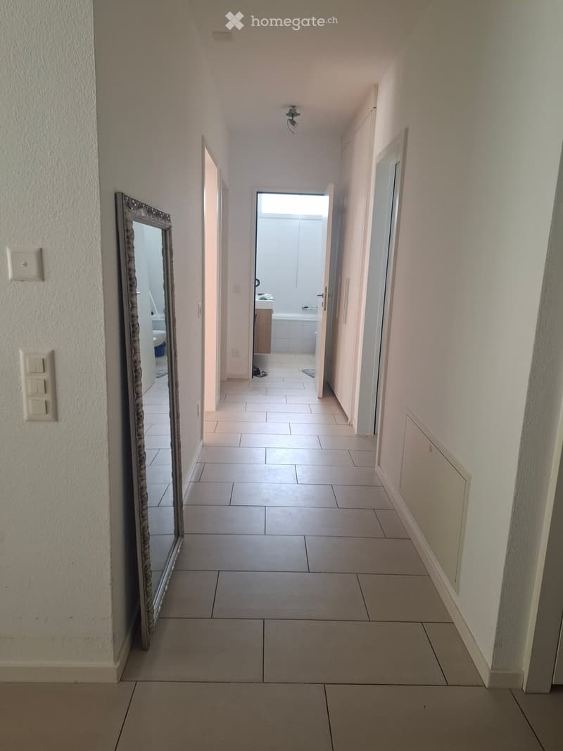 Empty hallway with tiled floor, white walls, and a large mirror on the left side. The hallway leads to a bathroom with a bathtub and a shower area visible at the end.