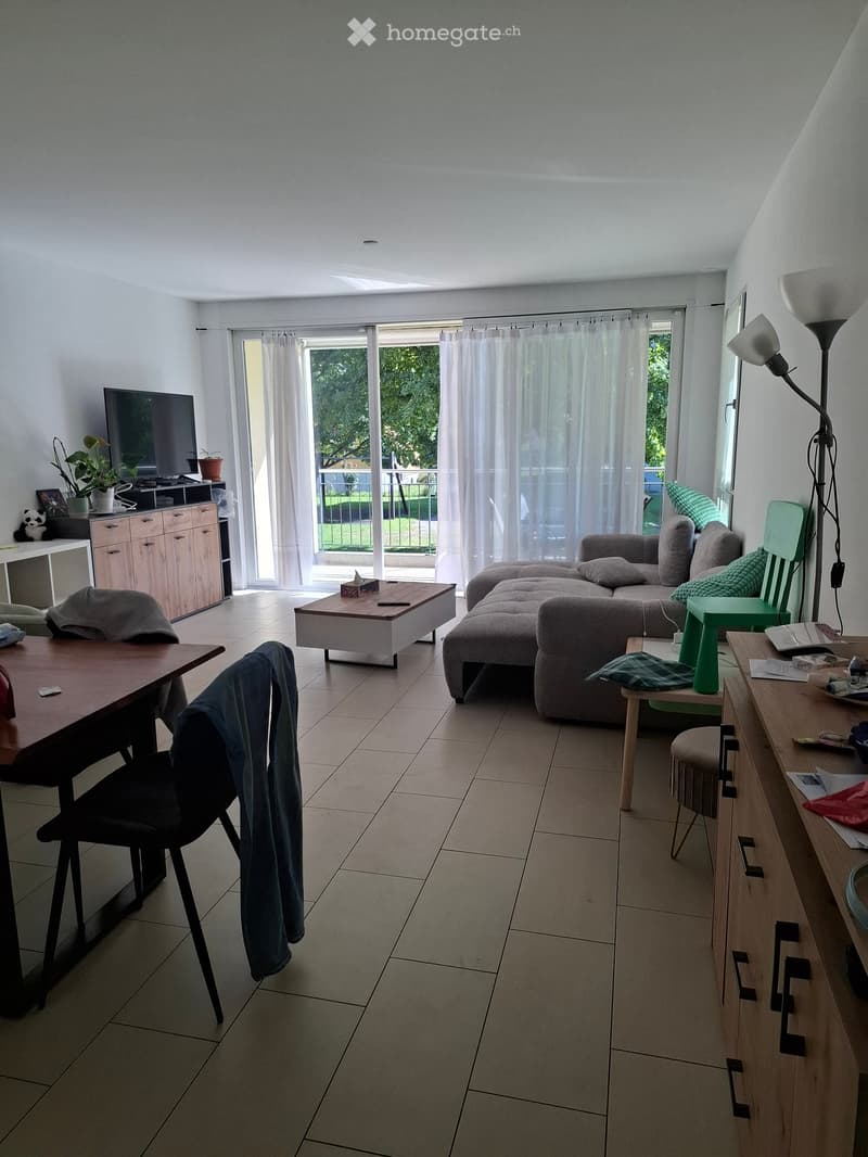 Modern living room with a grey sofa, coffee table, dining table, chairs, and a TV. Features large windows with a view to a balcony or terrace area.