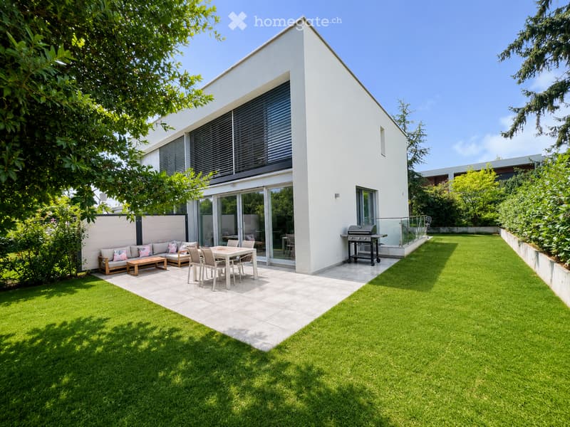 Modern outdoor space with a well-maintained lawn, patio area, and a contemporary house with large windows and a sliding glass door. Features include a sofa, coffee table, dining table with chairs, and a grill.