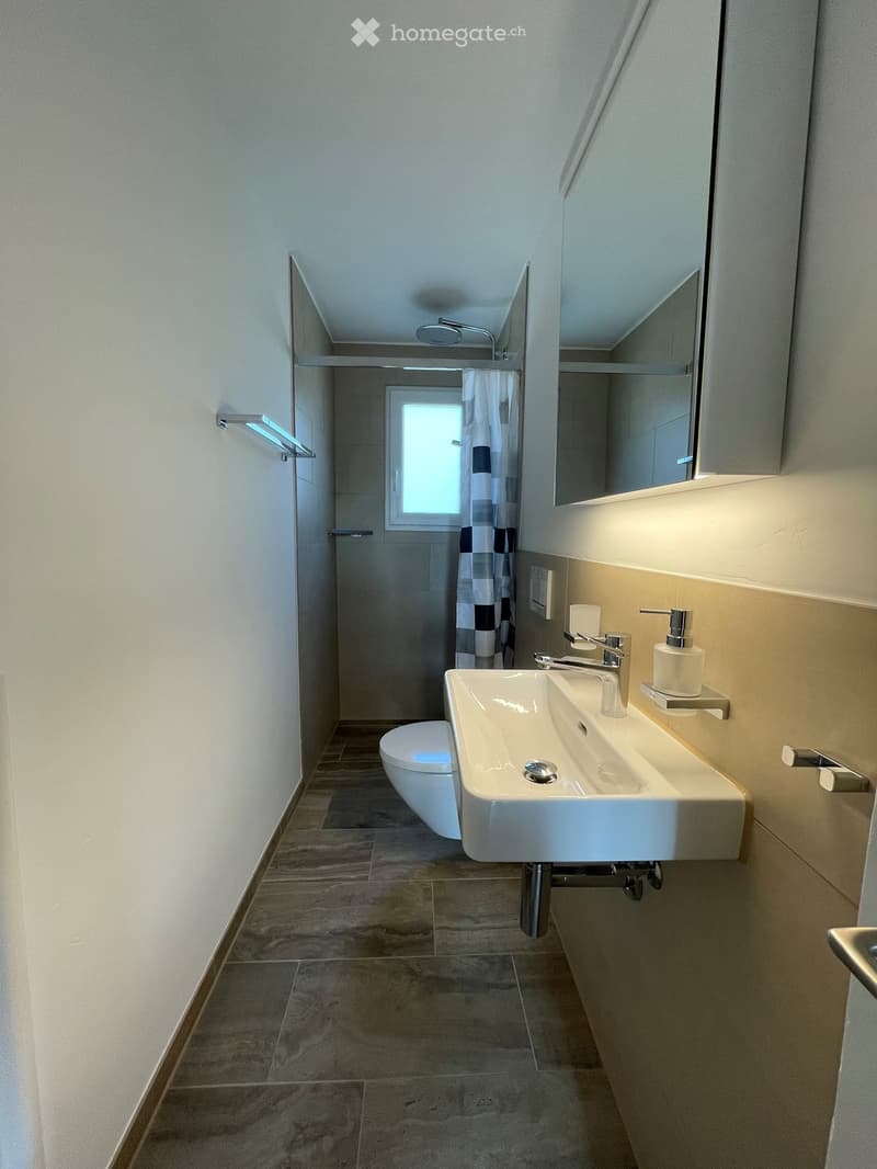 Modern bathroom with a white sink, toilet, large mirror, and shower area with a curtain. The space features tiled floors and walls with warm lighting.