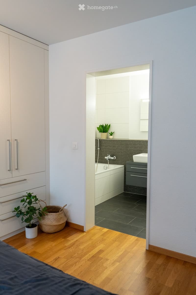 Modern bathroom with a bathtub, sink, and grey cabinets. The room features a dark tiled floor and a small plant on the windowsill.