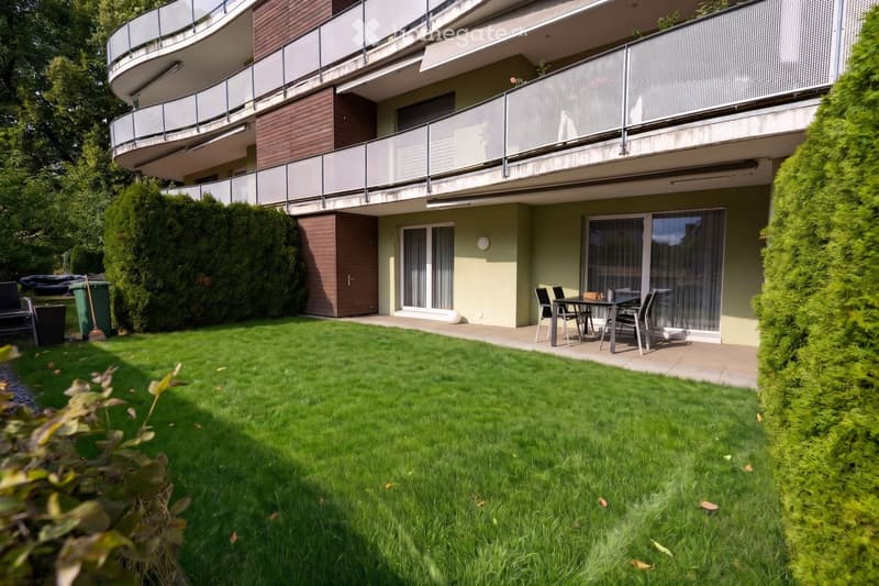 Well-maintained garden with green lawn, patio seating area, and modern apartment building with balconies in the background.