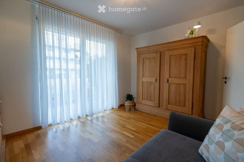 A modern living room with wooden flooring, a grey sofa, a wooden cabinet, a small table lamp, and a potted plant near the window with sheer white curtains.