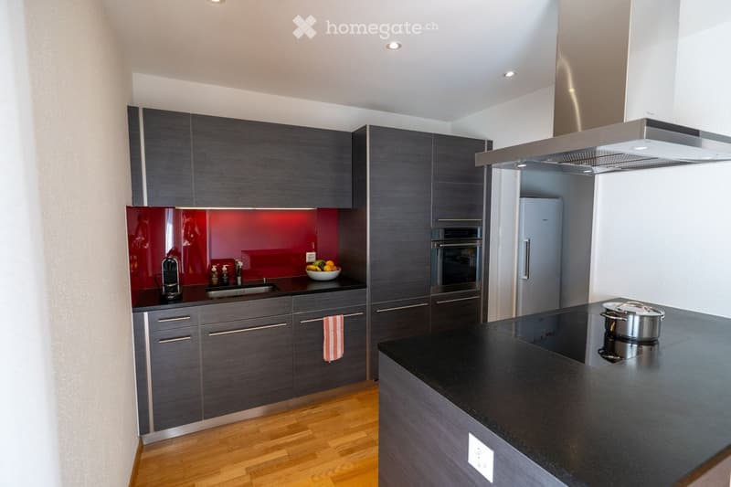 Modern kitchen with dark grey cabinets, a red backsplash, and stainless steel appliances including an oven, refrigerator, and coffee maker. The countertop features a bowl of fruit and a pot.