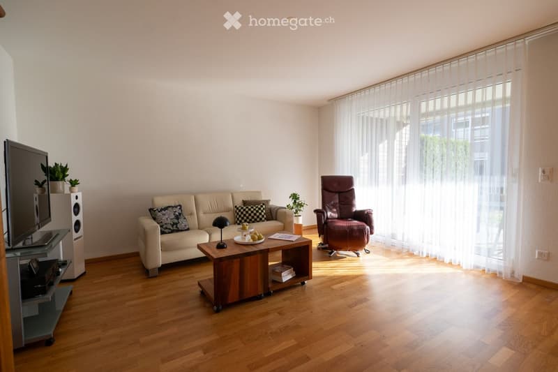 Bright living room with wooden flooring, large windows with sheer curtains, a beige sofa with patterned cushions, a dark red armchair, a wooden coffee table with a fruit bowl, a TV stand with a flat-screen TV, and a speaker system.