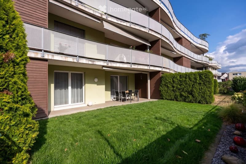 Well-maintained garden with green grass, trimmed hedges, and a small patio area with a table and chairs. The building in the background features balconies and modern architecture.