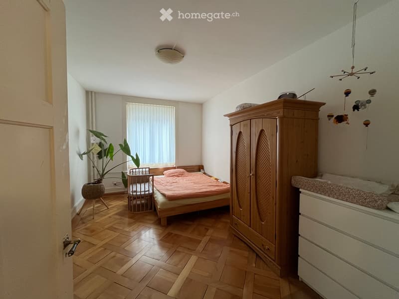 Cozy bedroom with wooden flooring, a double bed with pink bedding, a wooden wardrobe, a baby crib, a small plant, and a baby chair. A toy mobile hangs above the crib.
