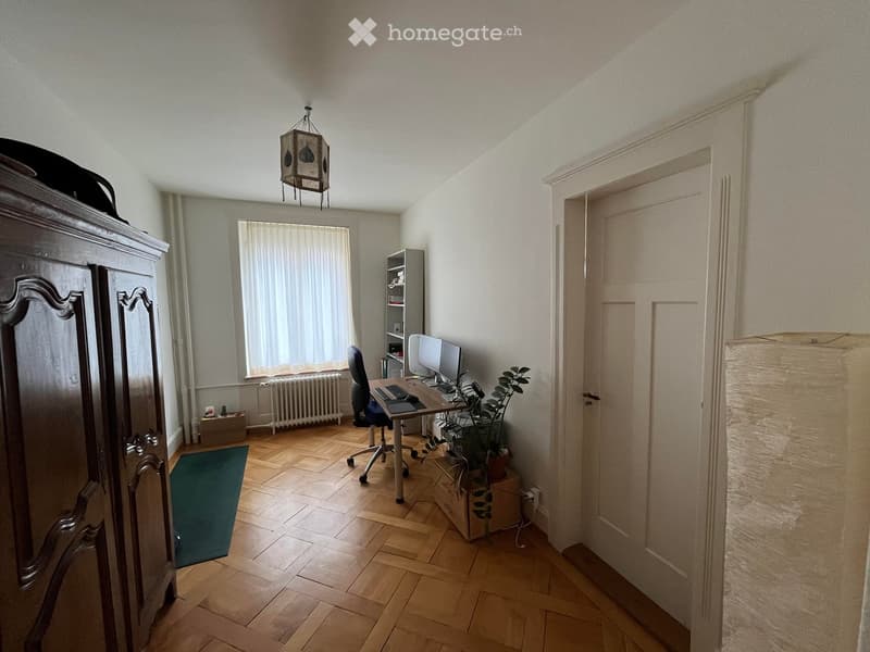 A well-lit room with wooden flooring, a large window with curtains, a desk with a computer and chair, a cabinet, and a small plant on the floor.