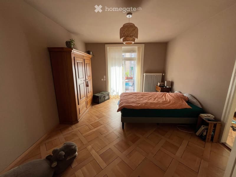 Cozy bedroom with wooden flooring, a bed with a pink blanket, a large wooden wardrobe, a nightstand, a radiator, and a small table with a chair near the window.