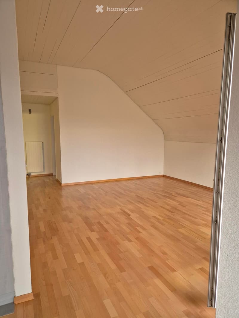 Empty room with light wooden flooring, white walls, and a slanted ceiling. There is a doorway leading to another room with a radiator visible.