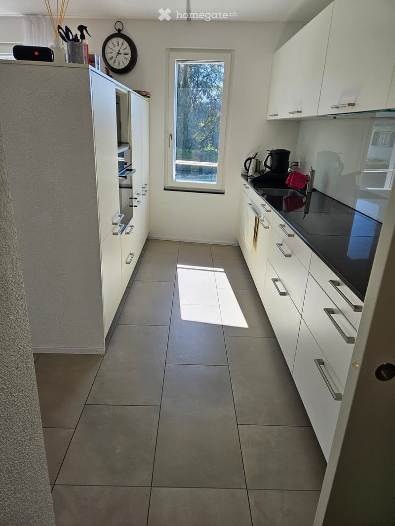 Modern kitchen with white cabinets, stainless steel handles, and a black countertop. Features a window, a clock, and various kitchen items on the counter.