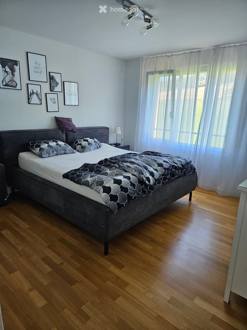 Modern bedroom with a large bed, grey headboard, patterned bedding, nightstand with lamp, and several picture frames on the wall. Wooden flooring and large window with sheer curtains.