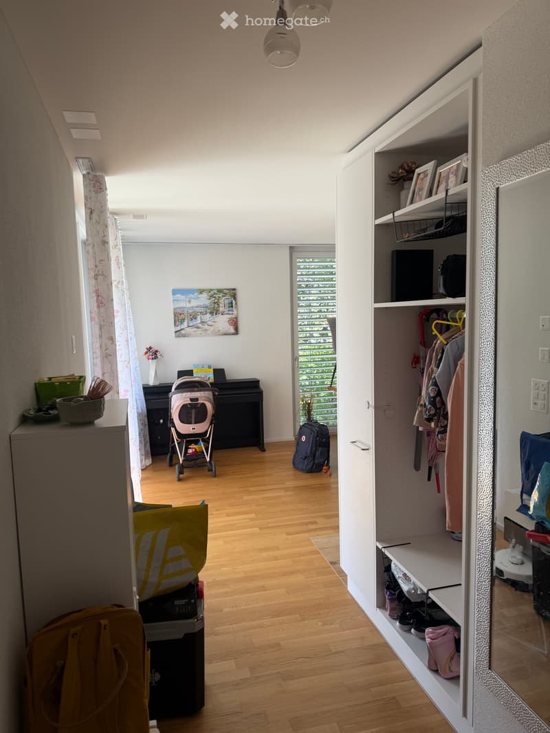 A well-lit hallway with wooden flooring, a piano, a baby stroller, and an open closet with clothes and shoes.