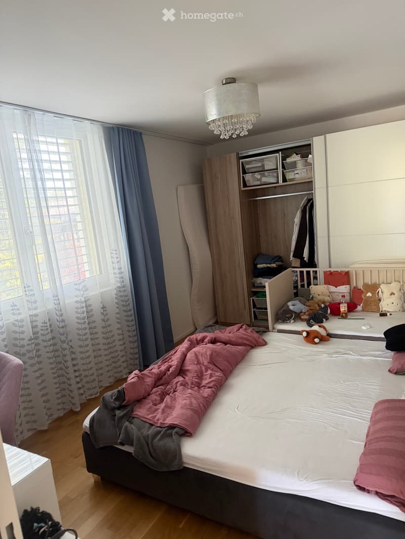 A well-lit bedroom with a large bed, a wardrobe, and a window with curtains. The bed is partially made with blankets and pillows, and there are toys on a changing table nearby.