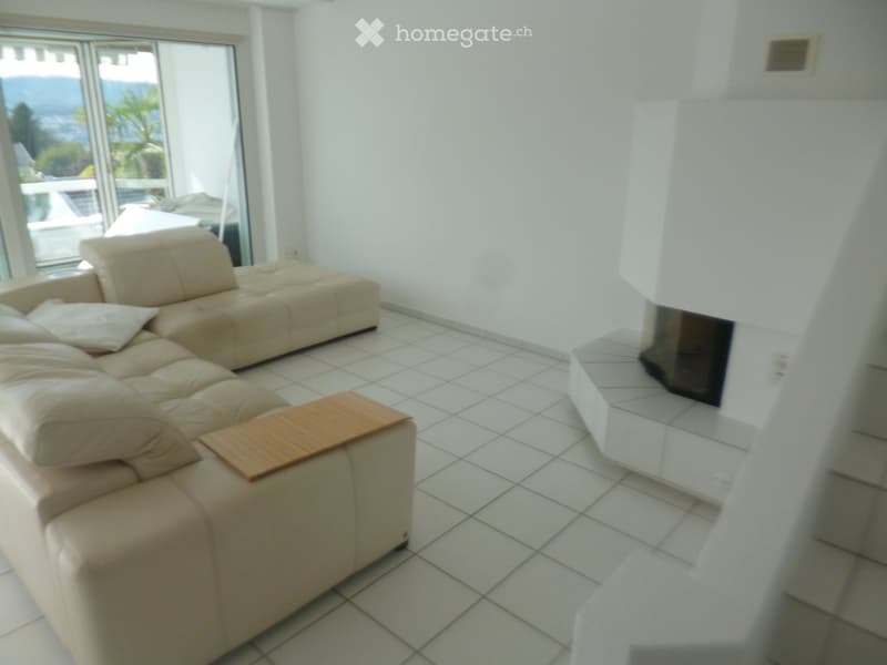 Bright living room with white tiled flooring, a beige sectional sofa, a wooden coffee table, and a fireplace, with large windows offering a scenic view.