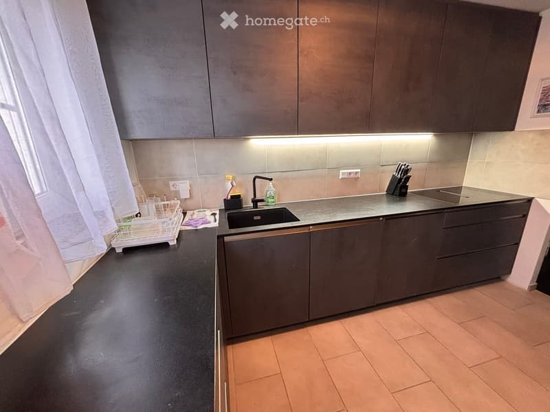Modern kitchen with dark grey cabinets, a stainless steel sink, and a countertop with various items including a dish rack, knife block, and a bottle. The floor is tiled in a warm beige color.