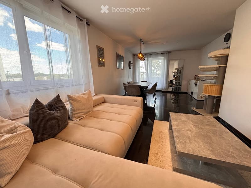 Modern living room with a white sectional sofa, coffee table, dining area with chairs, and a small kitchen area visible in the background.