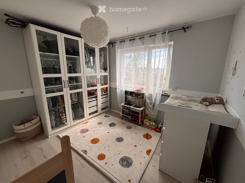 A well-lit bedroom with a large wardrobe, a loft bed, a small table with toys, and a colorful rug on the floor.