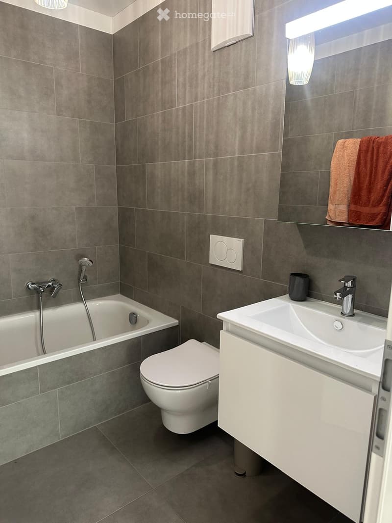 Modern bathroom with grey tiled walls, a bathtub, toilet, sink, and a mirror with a built-in light. A towel is draped over the mirror, and a cup sits on the sink counter.