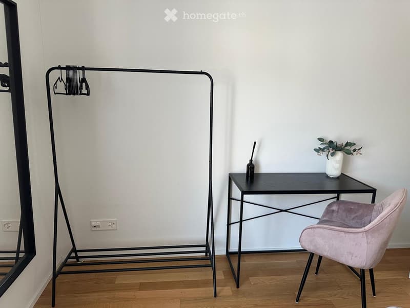 A small, modern room with a wooden floor, a black metal clothing rack, a small black table with a vase and bottle, and a pink upholstered chair.