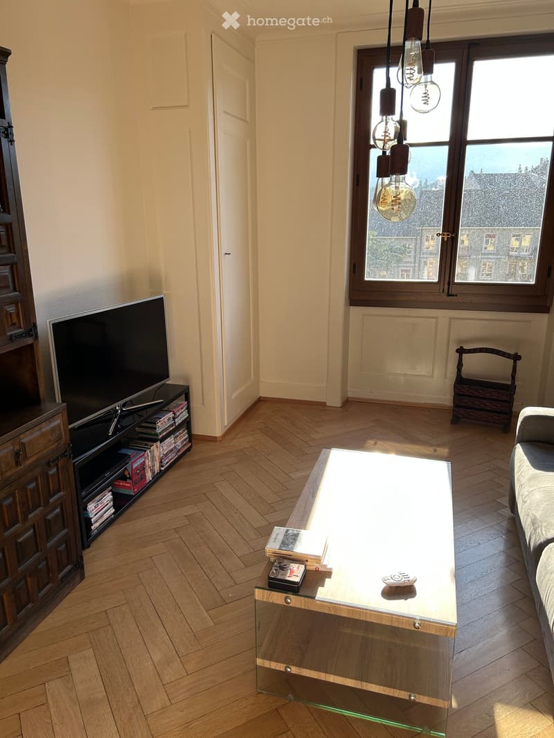 Living room with herringbone wooden flooring, large windows with city view, modern glass coffee table, sofa, TV on stand, and books on cabinet.