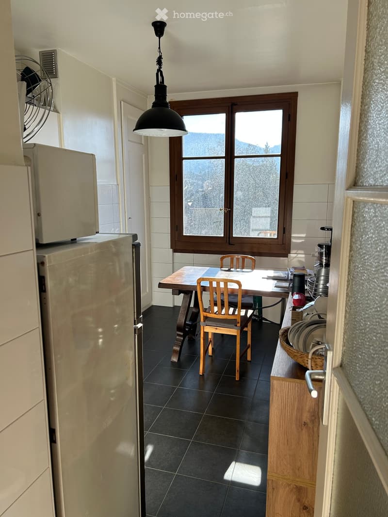 A modern kitchen with dark tiled flooring, a wooden dining table with chairs, a refrigerator, and a window offering a scenic mountain view.