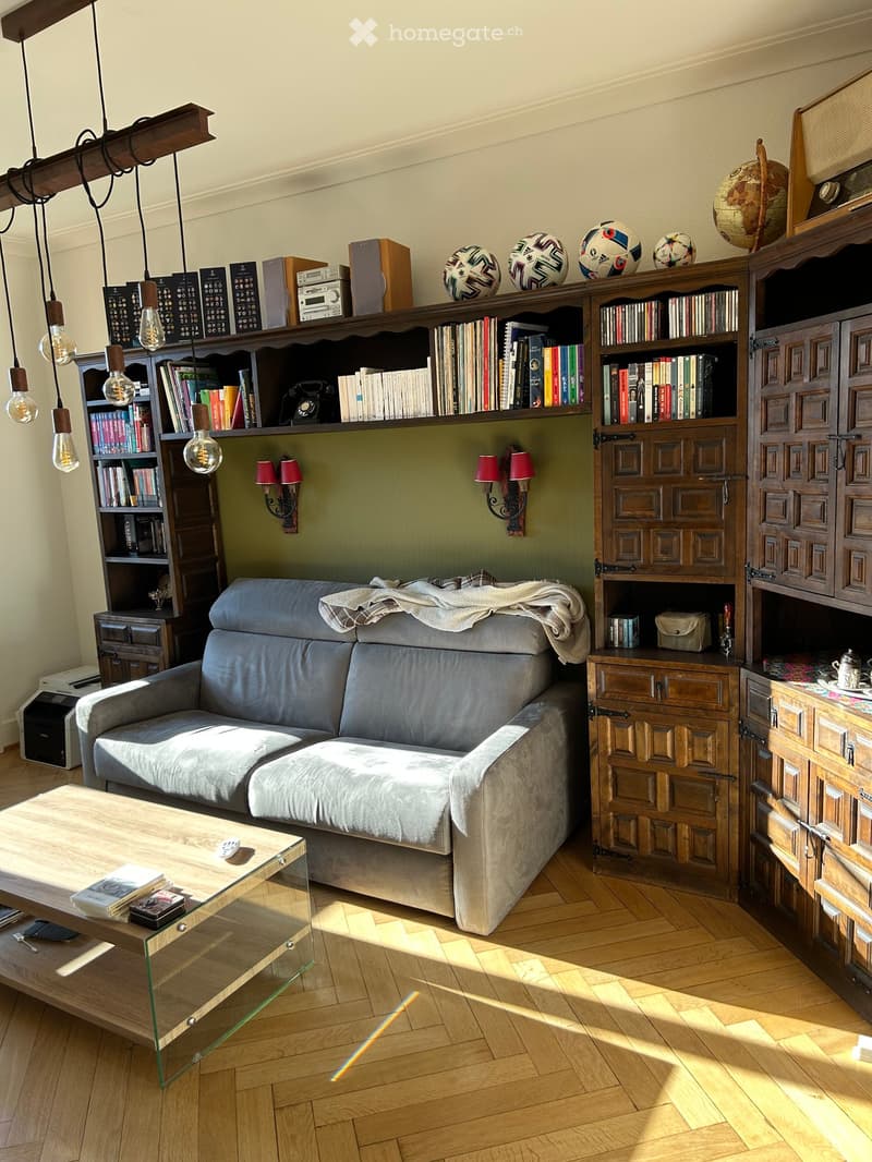Living room with a grey sofa, wooden coffee table, bookshelf filled with books and decorative items, globe, and wall-mounted lamps.