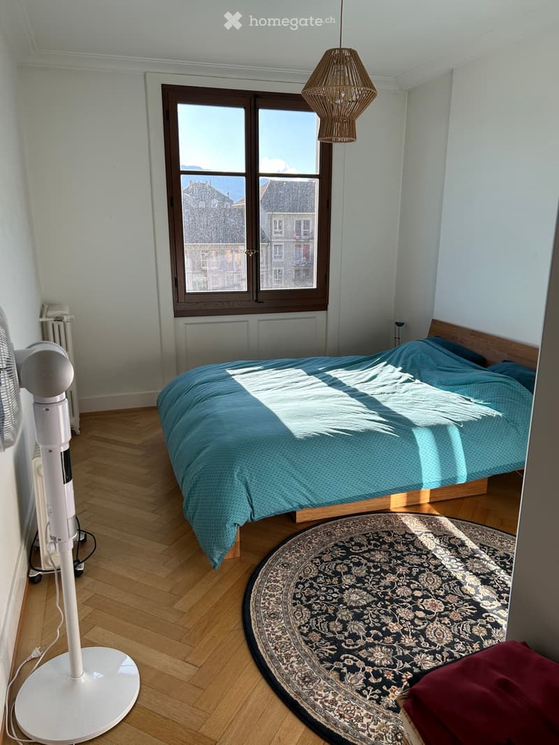Bright bedroom with wooden flooring, large window showing cityscape, bed with blue duvet, round patterned rug, standing fan, radiator, and hanging woven lamp.