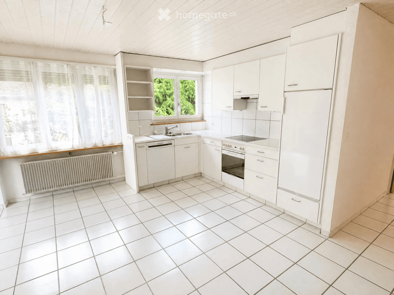 Bright kitchen with white tiled floor and walls, white cabinets, a refrigerator, oven, dishwasher, sink, and a window with white curtains.