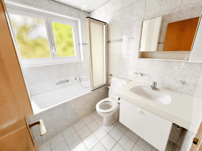 Modern bathroom with white tiles, a bathtub, toilet, sink, and a mirrored cabinet.