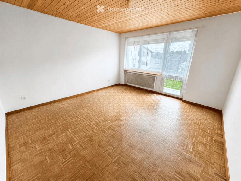 Empty room with wooden herringbone flooring, white walls, and large window with blinds. Radiator visible beneath window.