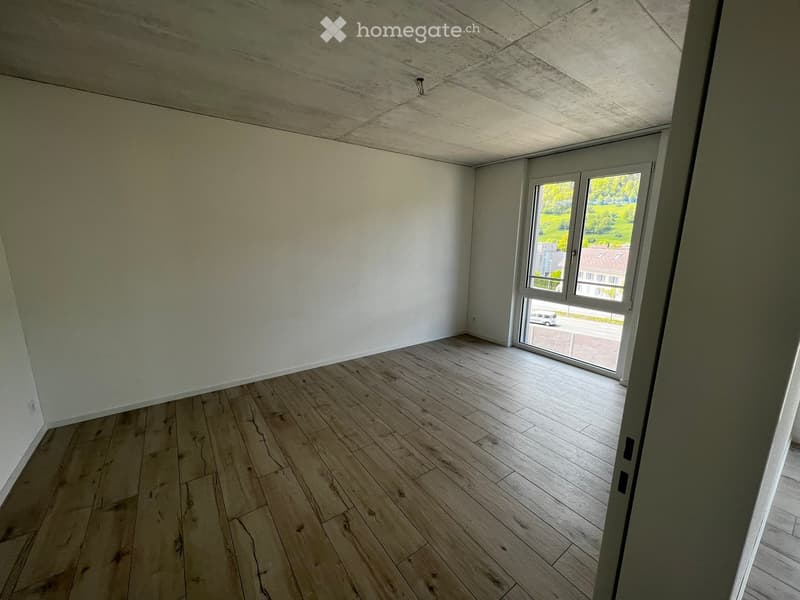 Empty room with wooden flooring, white walls, and a large window offering a view of the outside area.