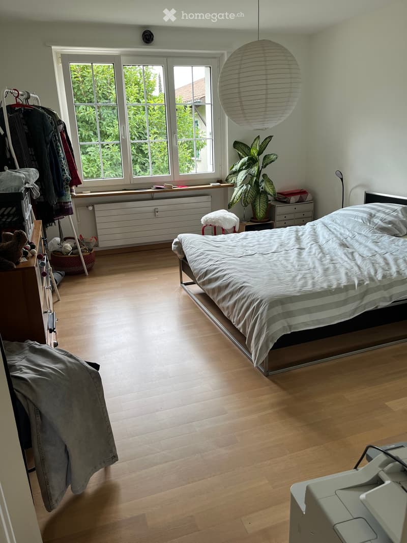 Bright bedroom with wooden flooring, large window, bed, small table with plant, stool, and wardrobe with clothes.