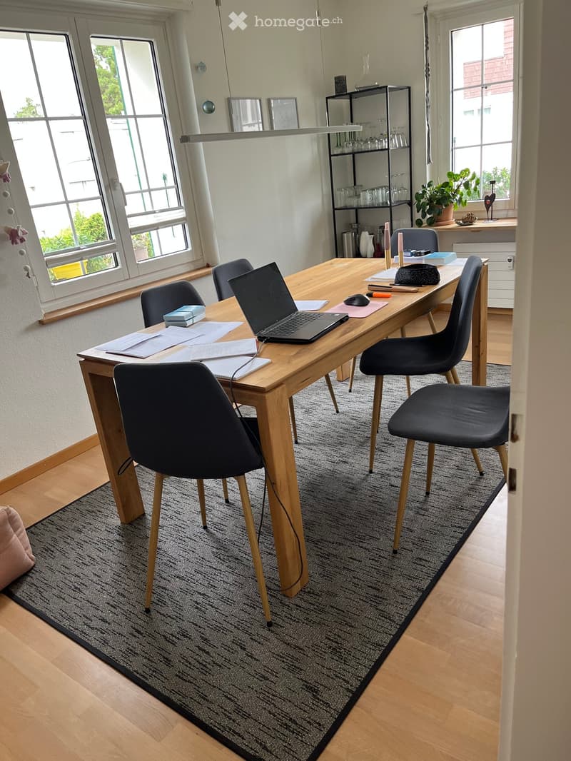 A modern, well-lit room with a wooden table, laptop, chairs, a shelf with glassware, and a small plant on the windowsill.
