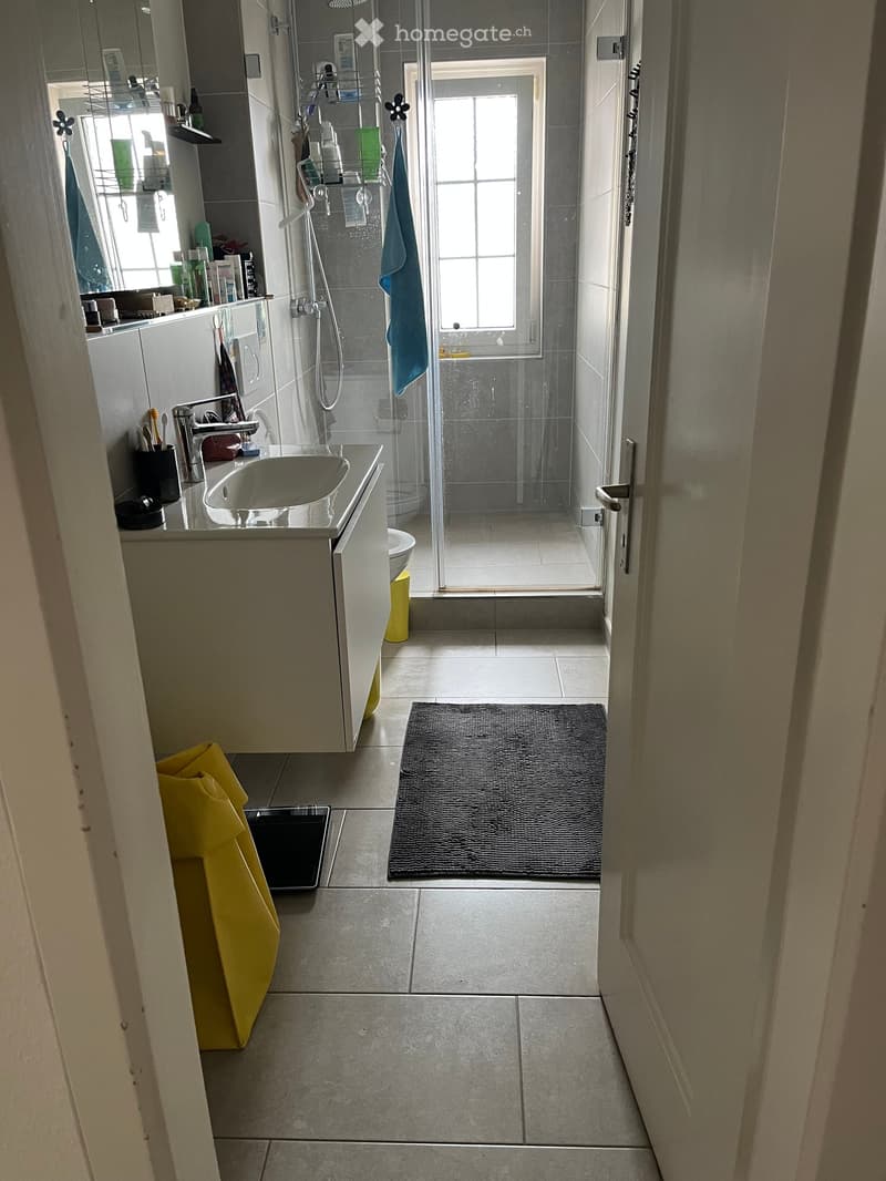 Modern bathroom with a sink, mirror, shower, and toilet. Features a grey tiled floor, a black bathroom rug, and a yellow towel on the side.