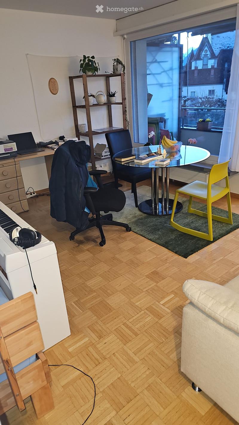 A modern, well-lit room with wooden herringbone flooring, a large window with a view, a sofa, a round glass table with chairs, a desk with a laptop, and a shelf with decorative items.