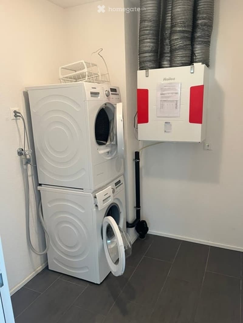 Compact bathroom with stacked white washing machine and dryer, dark tiled floor, white walls, and a water heater unit mounted on the wall.