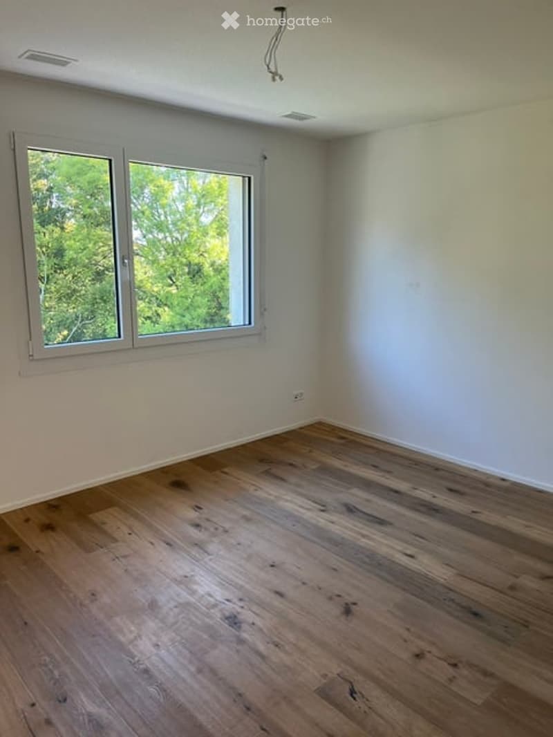 Empty room with wooden flooring and large windows showing greenery outside. No furniture or equipment present.