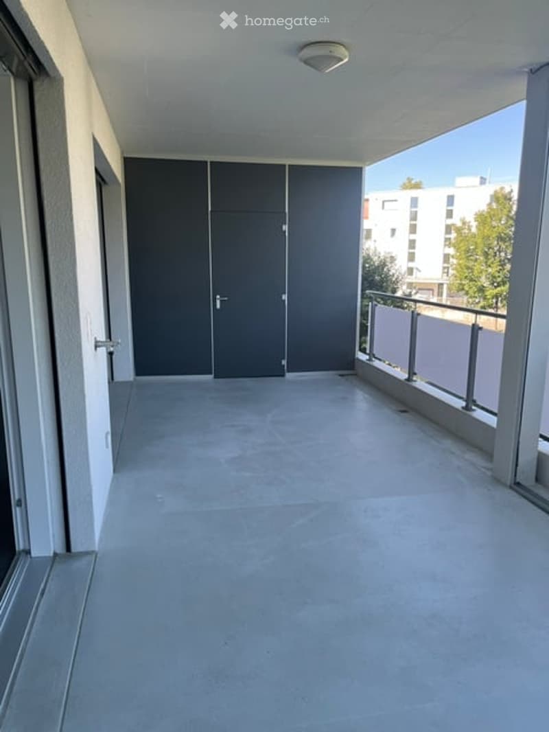 Modern balcony with grey sliding doors, concrete floor, metal railing, and a view of a building and trees.