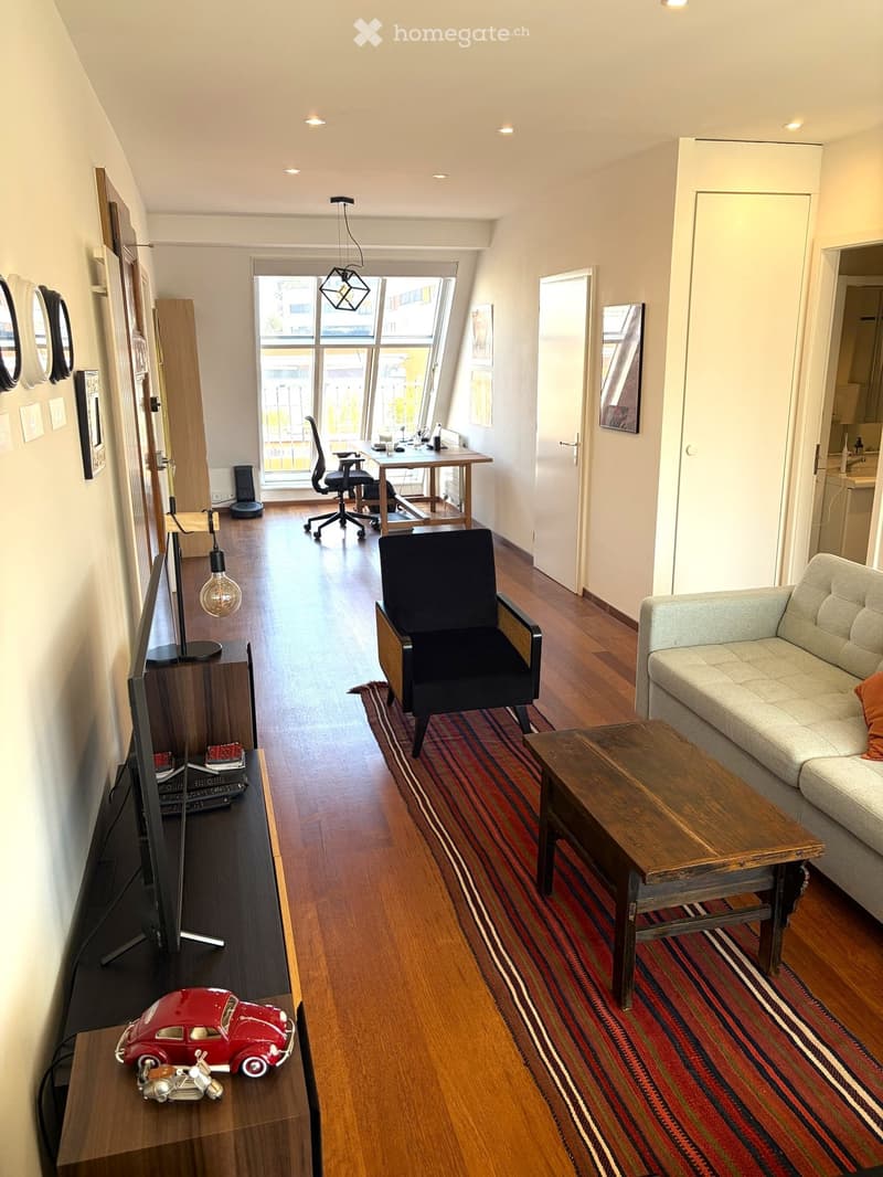 A modern, open-concept living and working space with wooden flooring, large windows, a sofa, coffee table, desk, and office chair.