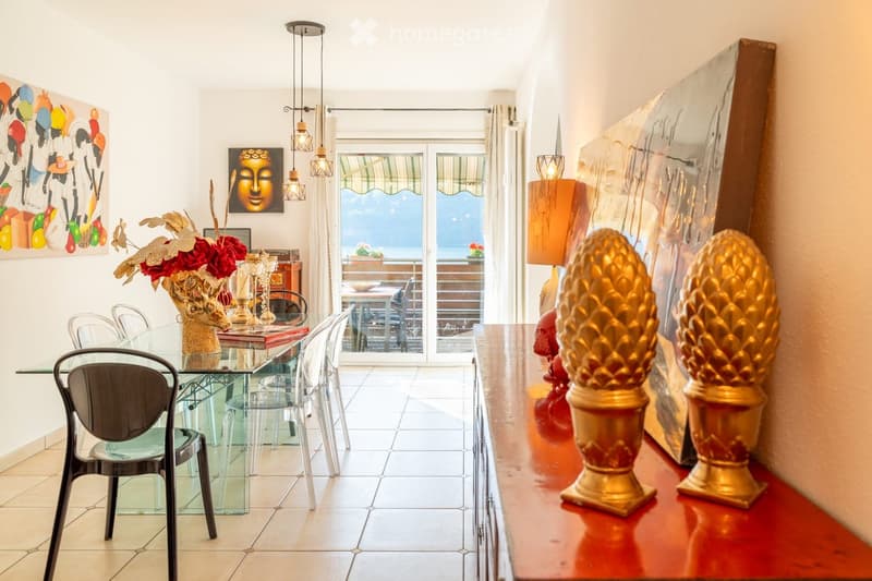Dining room with a glass table, modern chairs, decorative vases, candle holders, and artwork on the walls. Large windows offer a view of the outdoor area.