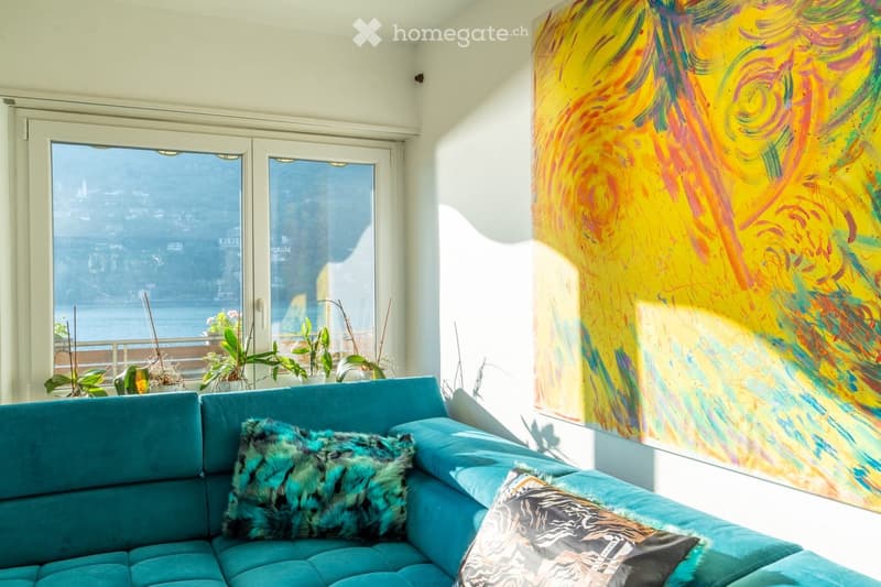 Bright living room with a teal sectional sofa, colorful abstract painting, and large windows offering a scenic view of the water.