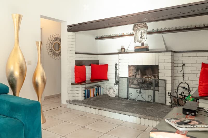 Living room with a brick fireplace, red cushions, a teal sofa, two tall gold vases, a coffee table with magazines, and decorative shelves with a Buddha head and small items.